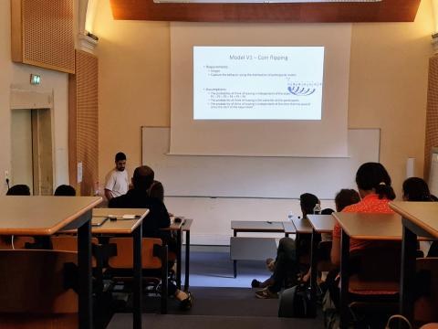 Daniel Yamin at the front of a lecture hall presenting a slide titled 'Model V1 — Coin flipping'